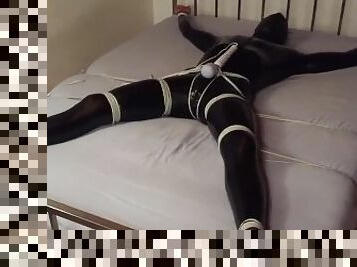 Male in bed bondage in a shiny catsuit with a leather hood.